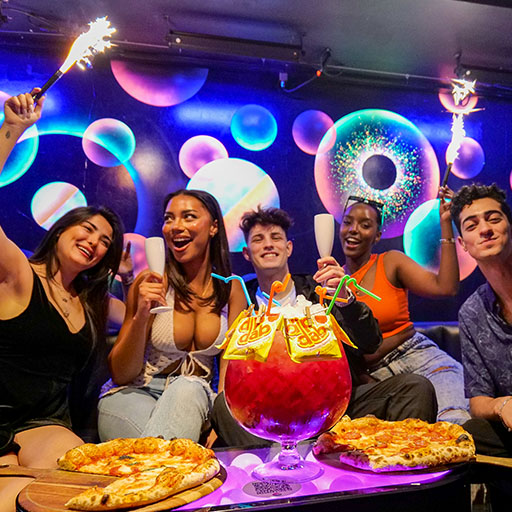 a group of friends with a giant sharing cocktail, drinking champagne, eating pizza and dancing with sparklers in their hands