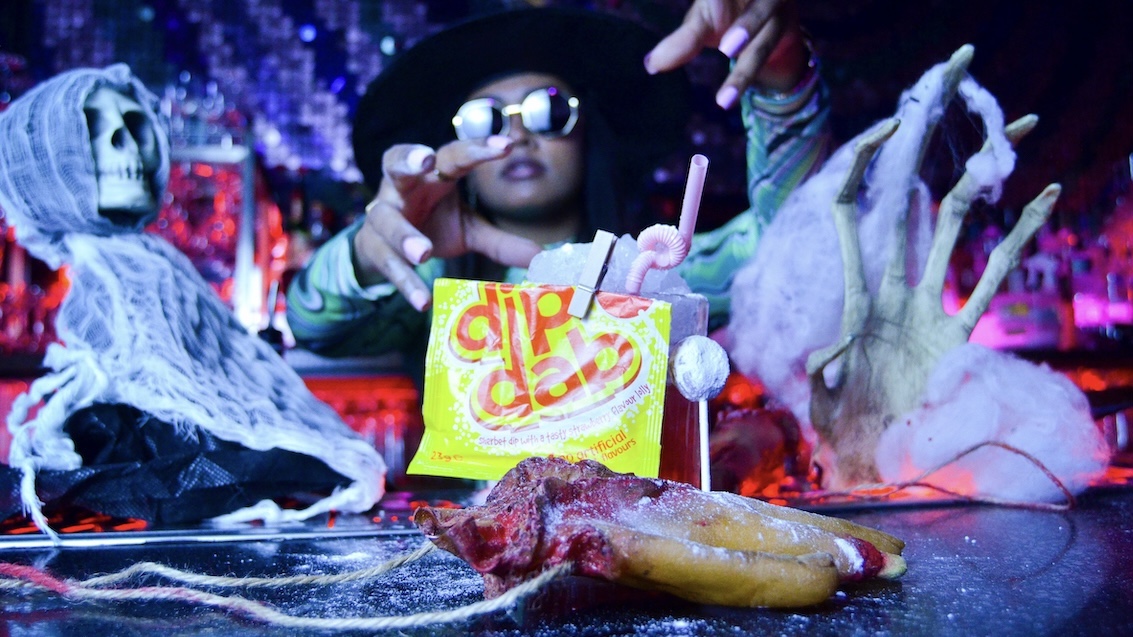 a halloween witch casting a spell on a cocktail with halloween decorations around her