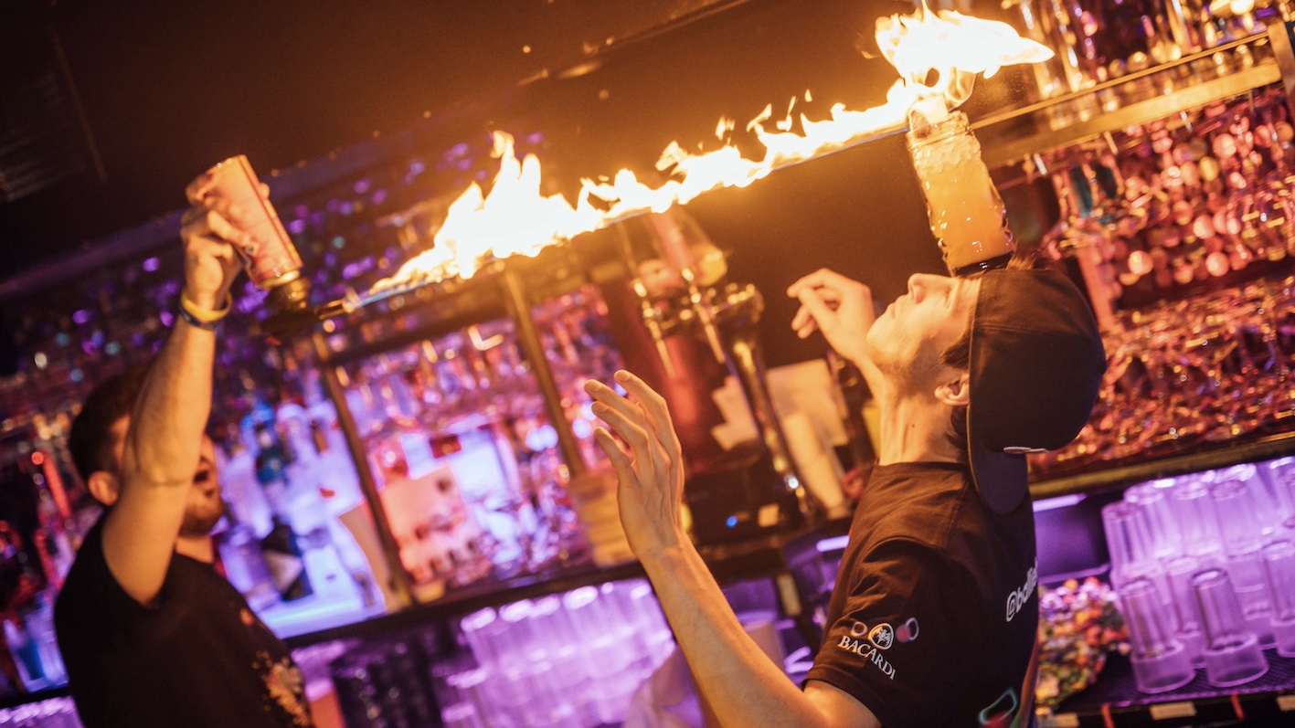 two bartenders, one has a cocktail balancing on his head, the other is pointing a flame at the cocktail, warming a marshmallow on the cocktail