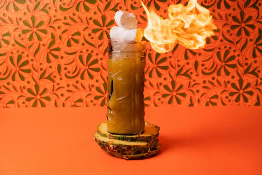 a cocktail standing on two slices of pineapple, the cocktail has a marshmallow on top which is being roasted by a big flame